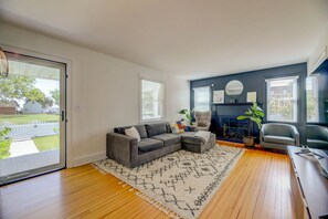 A living room with a grey sectional sofa, a white rug, a fireplace, and a large window with blinds. - Marina View Cambridge Cottage: House on Water (Cambridge)