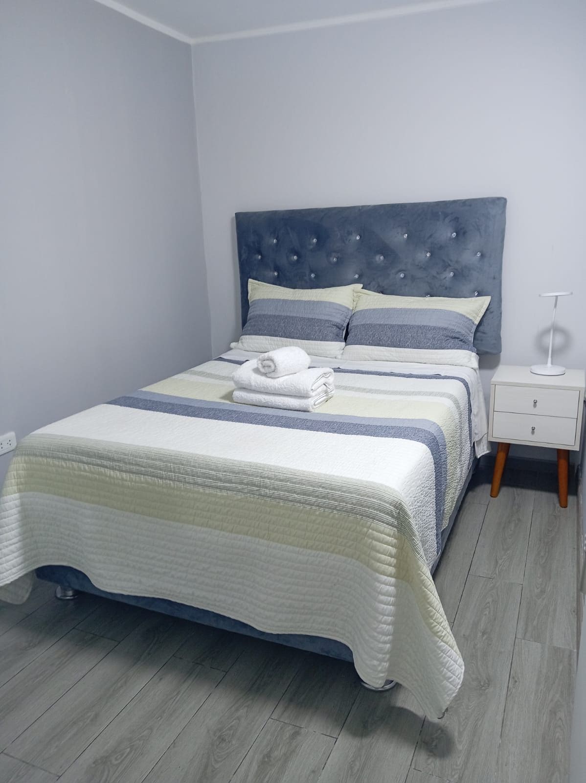 A neatly made bed with a grey headboard, a bedside table with a lamp, and a grey wooden floor.