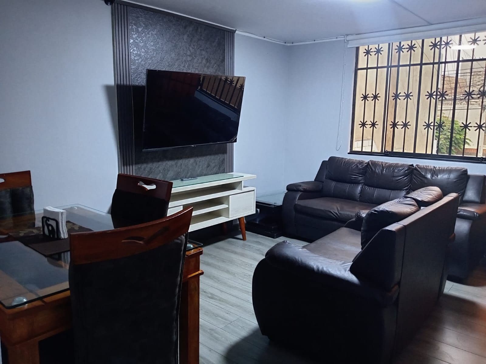 A living room with a flat-screen TV mounted on the wall, a leather sofa, a glass coffee table, and a wooden chair.