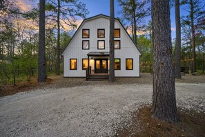 A two-story wooden cabin with a front porch, surrounded by trees. - Hot Tub + Game Room + Playground | Sleeps 14 (Broken Bow)