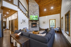 A spacious living room with a stone fireplace, a large gray sectional sofa, and a wooden ceiling. - Hot Tub + Game Room + Playground | Sleeps 14 (Broken Bow)