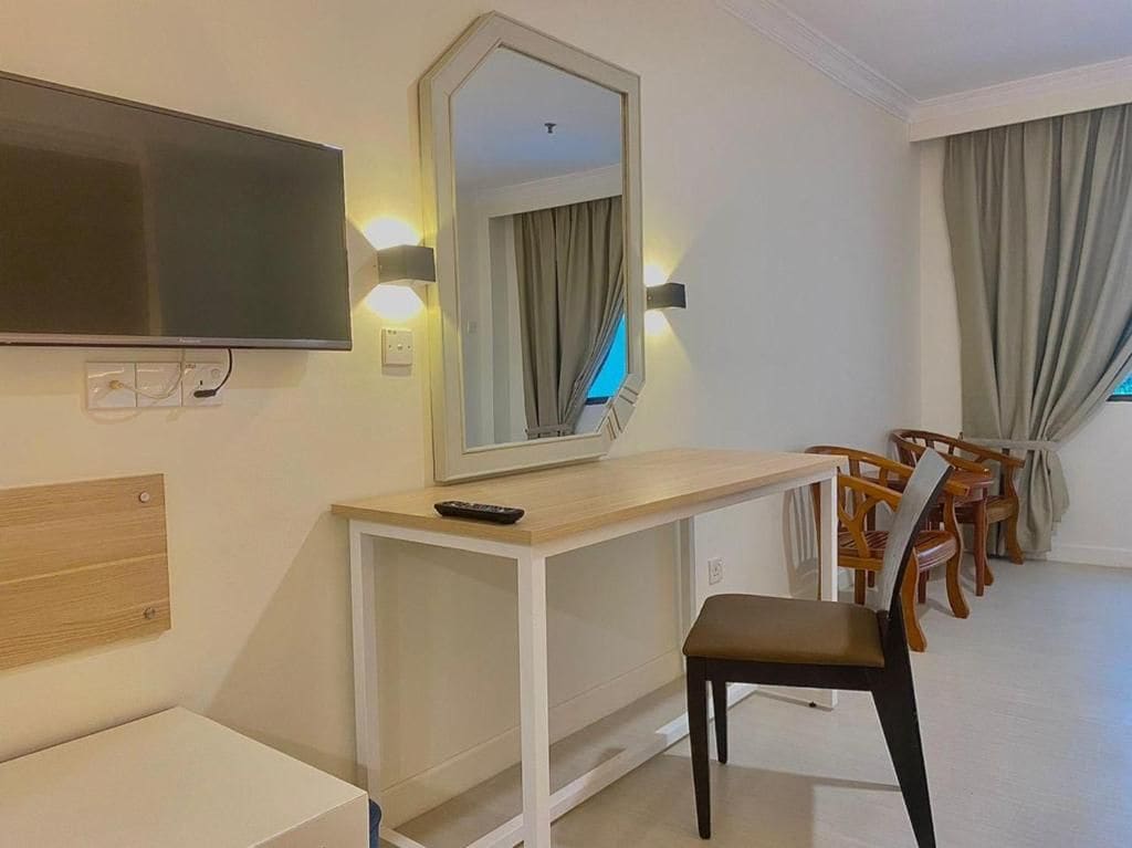A hotel room with a flat-screen TV mounted on the wall, a desk with a chair, and a large mirror.