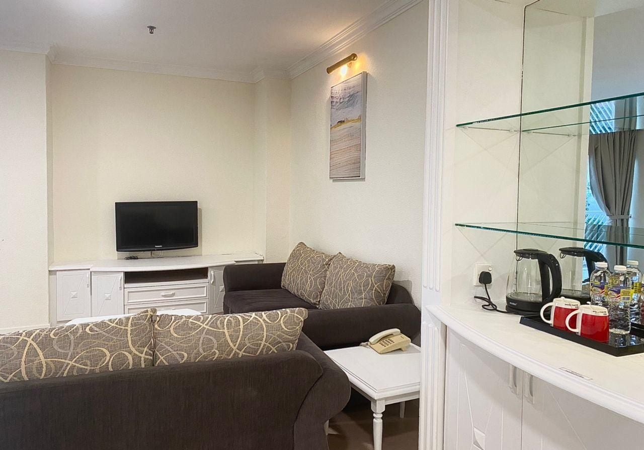 Habitación de hotel con sofá, televisión, una pequeña mesa con teléfono, mesa de vidrio con tetera y tazas, y una lámpara de pared.