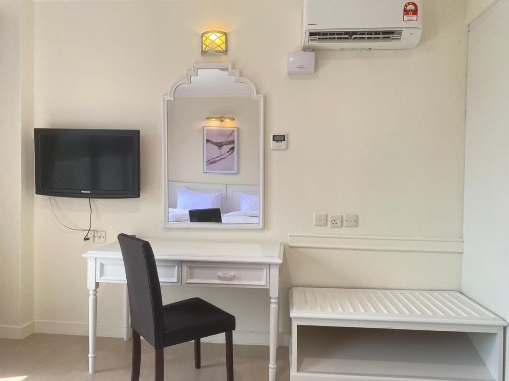 Una habitación con un escritorio blanco, una silla negra, un televisor de pantalla plana, aire acondicionado montado en la pared y un estante pequeño.