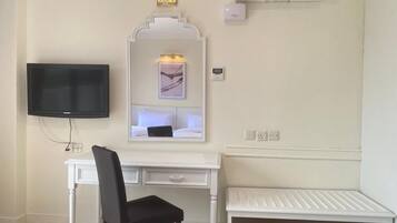 Una habitación con un escritorio blanco, una silla negra, un televisor de pantalla plana, aire acondicionado montado en la pared y un estante pequeño.