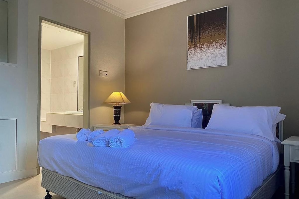 A neatly made bed with white linens and a folded towel, a bedside lamp, and a framed artwork on the wall.