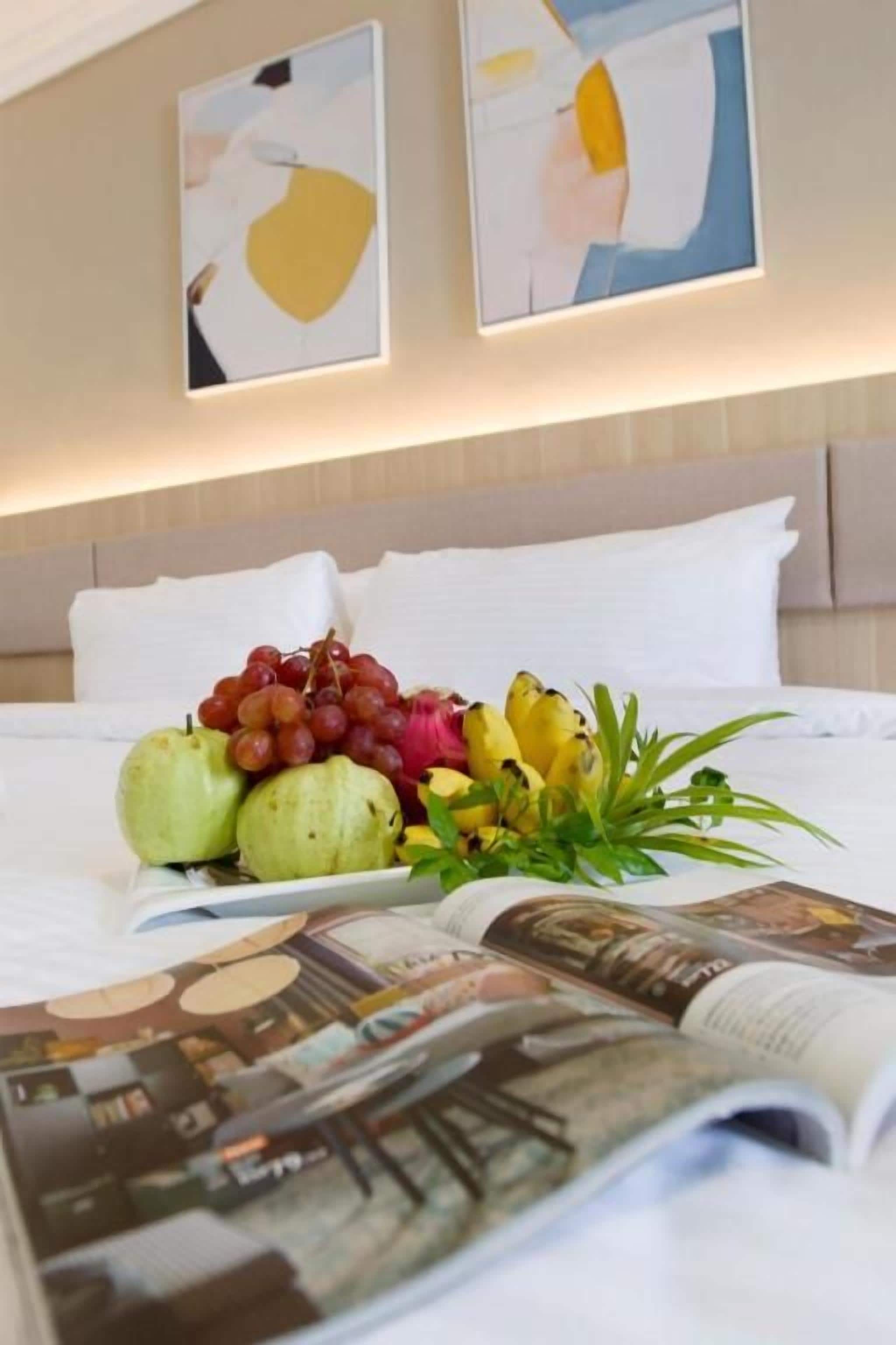 Quarto de hotel com uma cama, um prato de frutas na mesa de cabeceira e duas pinturas abstratas na parede.