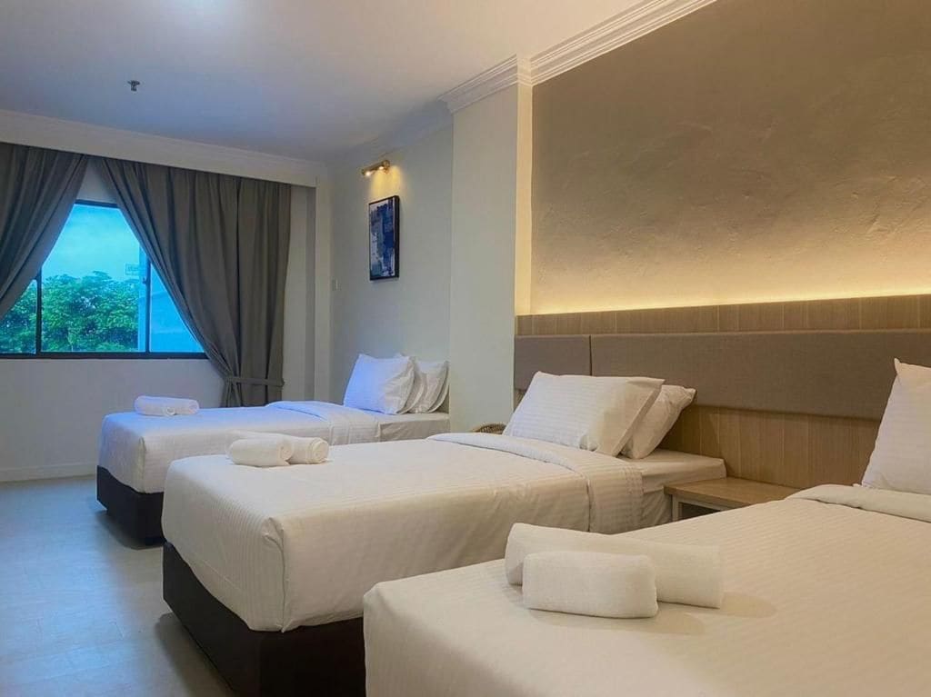 A hotel room with three beds, a large window with curtains, and a wall-mounted light.