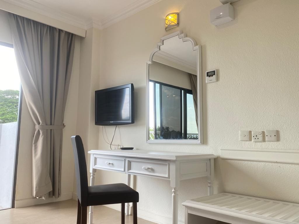 A room with a white desk, a black chair, a flat-screen TV, a large mirror, and a window with curtains.