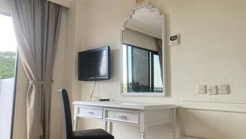 A room with a white desk, a black chair, a flat-screen TV, a large mirror, and a window with curtains.