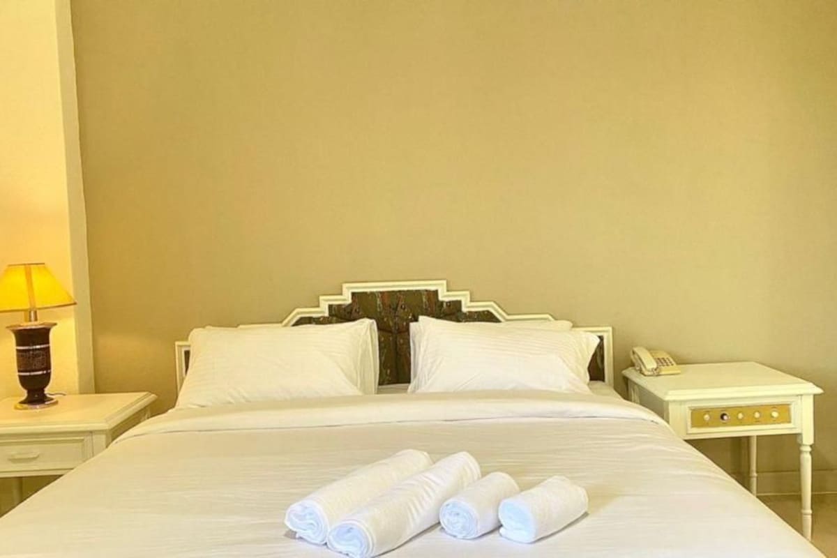 A neatly made bed with white linens and rolled towels, a bedside lamp, a telephone, and a small nightstand.