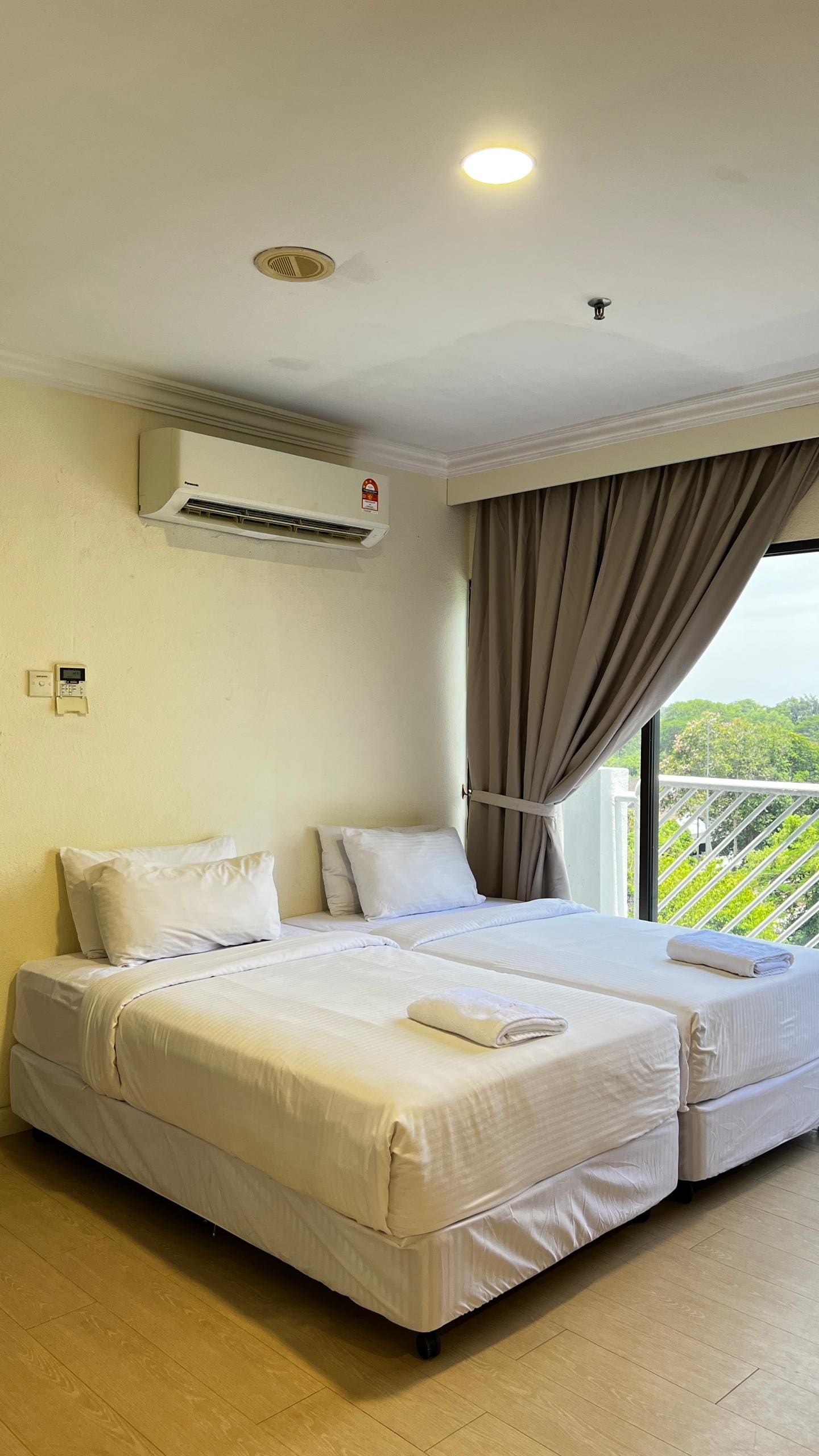 A hotel room with two beds, an air conditioning unit, a window with curtains, and a view of greenery.