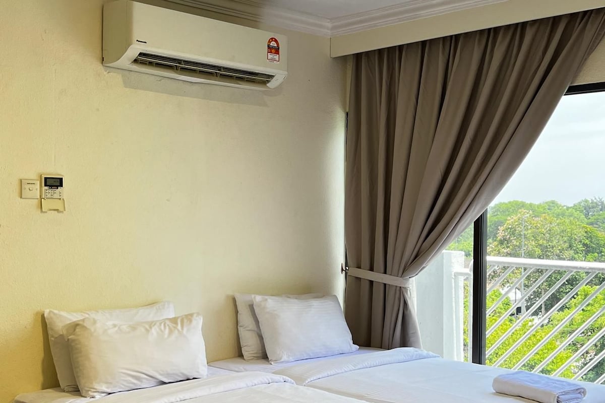 A hotel room with two beds, an air conditioning unit, a window with curtains, and a view of greenery.