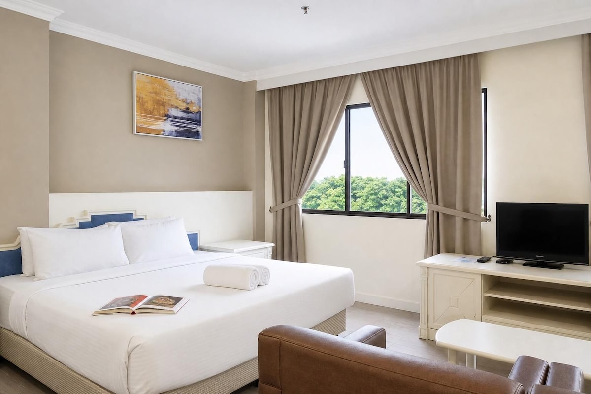 A hotel room with a bed, a desk with a television, a window with curtains, and a painting on the wall.