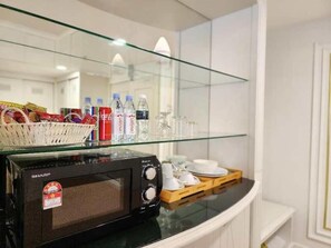 A microwave on a glass shelf with a basket of snacks, water bottles, and a tray with cups and a plate. - Swiss Avenue Hotel (Sungai Petani)