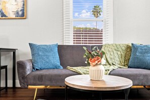 A living room with a grey sofa, blue cushions, a round wooden coffee table, and a vase with flowers. - Craftsman Bungalow in D'heart of the Arts District (Houston)