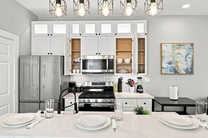 A modern kitchen with white cabinets, a stainless steel stove, and a microwave. There is a dining table set with plates and glasses. - Craftsman Bungalow in D'heart of the Arts District (Houston)