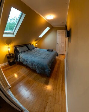 Quarto com claraboia, cama com colcha azul, abajur e porta.