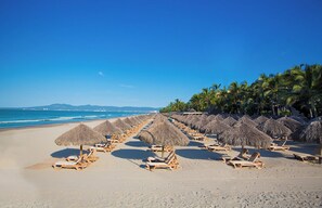 Una spiaggia con file di lettini e ombrelloni di paglia, palme e un cielo azzurro intenso.