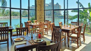En restaurant med store vinduer der giver udsigt over vand og bygninger. Der er dækket borde med stole, og drikkevarer samt tallerkener står på bordene.