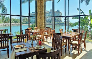 Un ristorante con ampie vetrate che si affacciano su un corpo d'acqua e degli edifici. I tavoli sono apparecchiati con sedie, e ci sono bevande e piatti sopra.