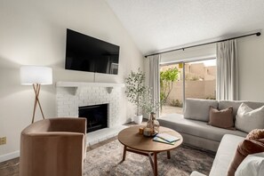 Uma sala de estar moderna com lareira, uma TV de tela plana acima dela, um sofá, uma mesa de centro com livros e uma vela, e um abajur.