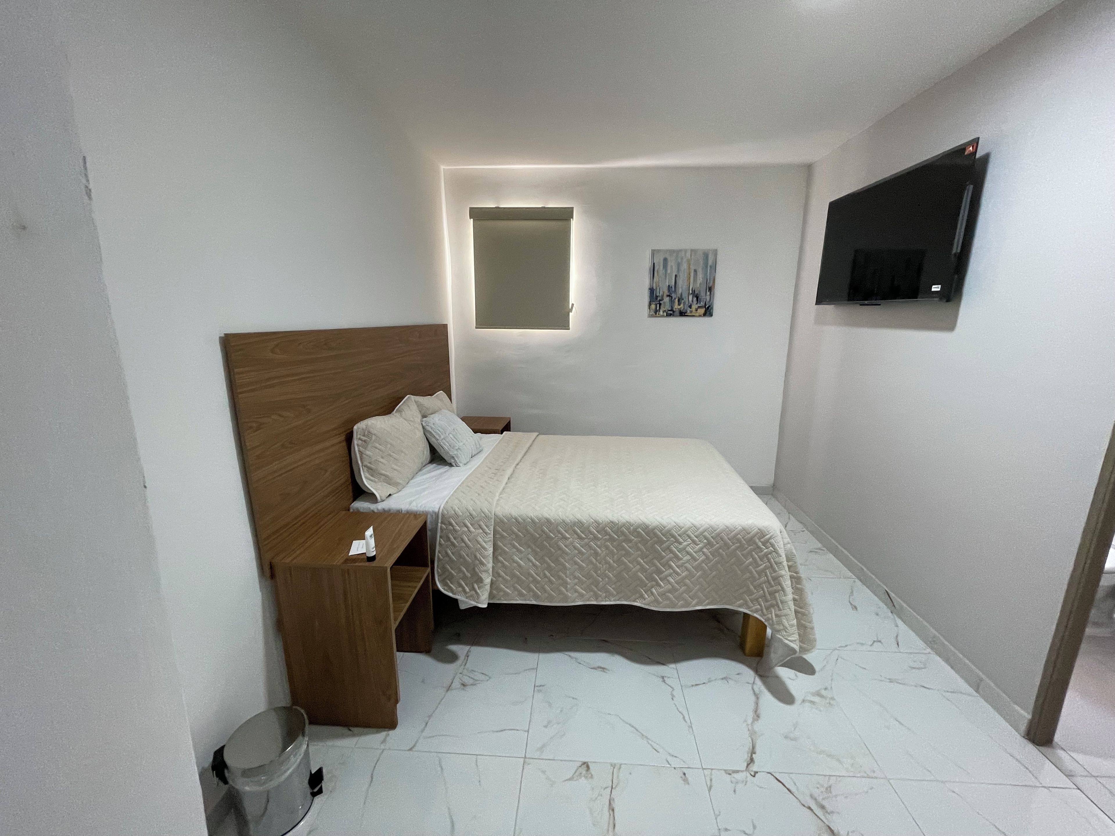 Un dormitorio pequeño y ordenado con una cama individual, una mesita de noche, un televisor montado en la pared y una ventana pequeña con persianas.