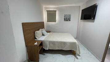 Un dormitorio pequeño y ordenado con una cama individual, una mesita de noche, un televisor montado en la pared y una ventana pequeña con persianas.