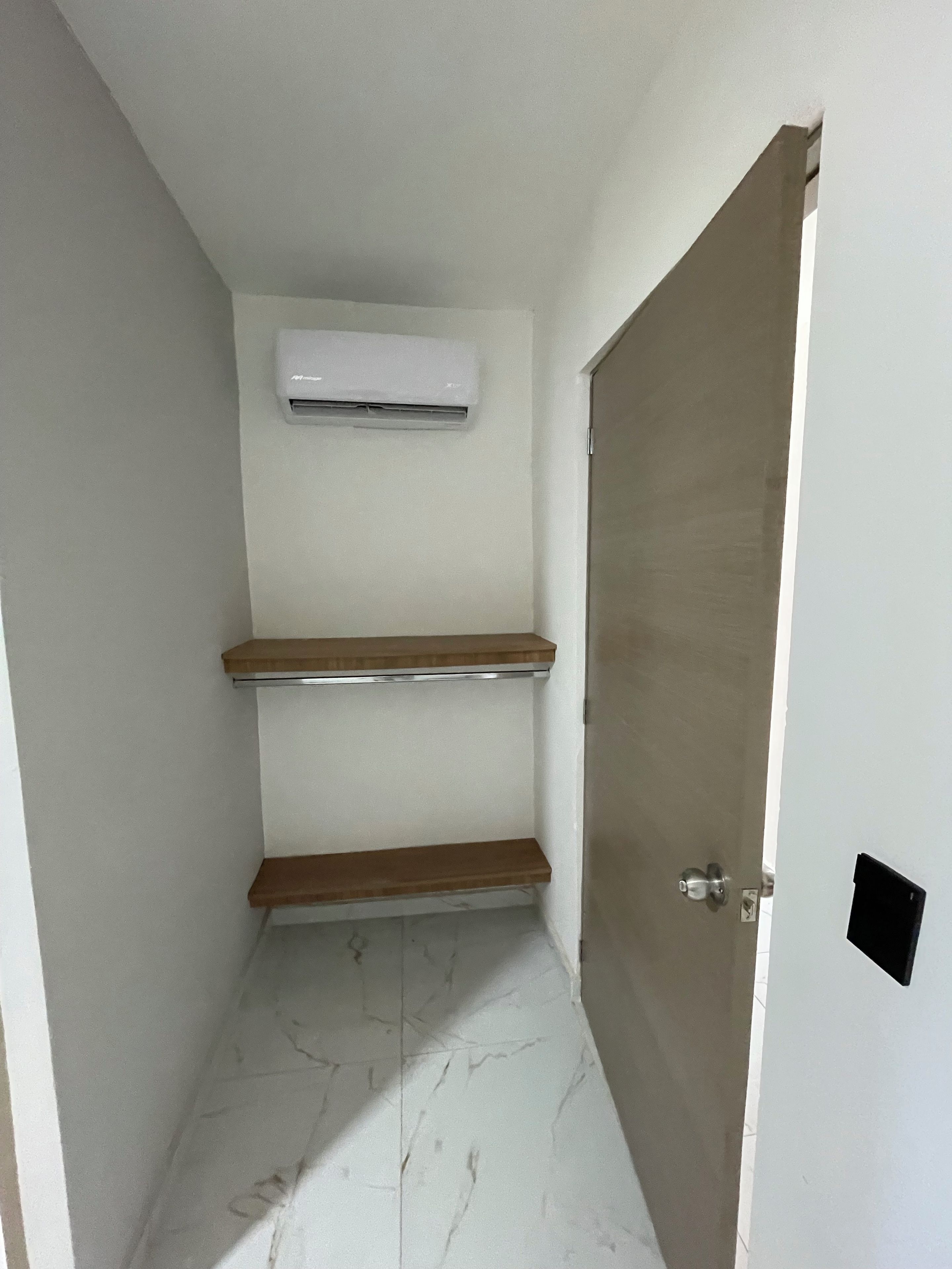 Um corredor estreito com uma porta à direita, um aparelho de ar condicionado branco na parede e duas prateleiras de madeira.
