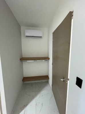 A narrow hallway with a door on the right, a white air conditioning unit on the wall, and two wooden shelves.