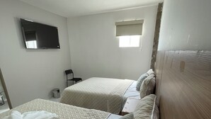 Et hotelværelse med to senge, et fladskærms-tv på væggen, et lille vindue med persienne og en stol.