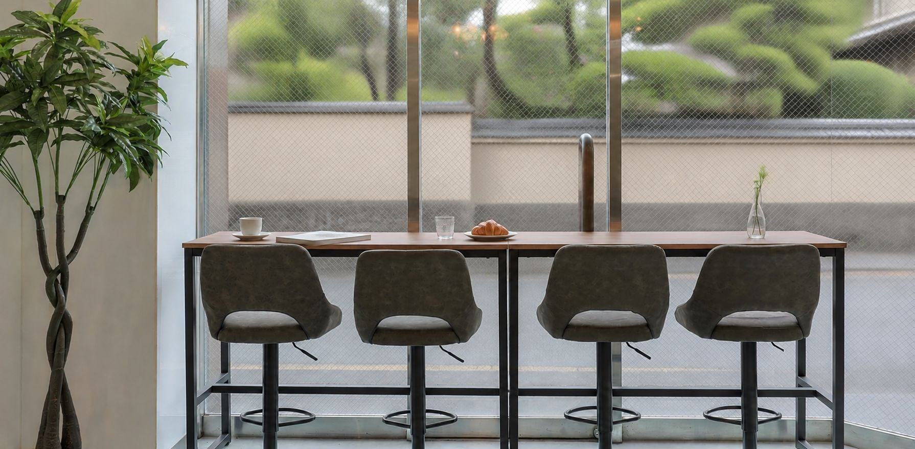 A modern interior with a wooden table and four grey chairs, a potted plant, and a view of greenery outside.