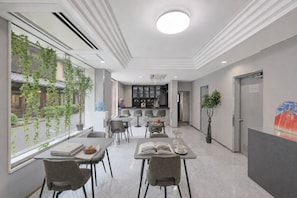 Un hall d'hôtel moderne avec un espace repas, une cuisine en arrière-plan et une grande fenêtre donnant sur un espace verdoyant.