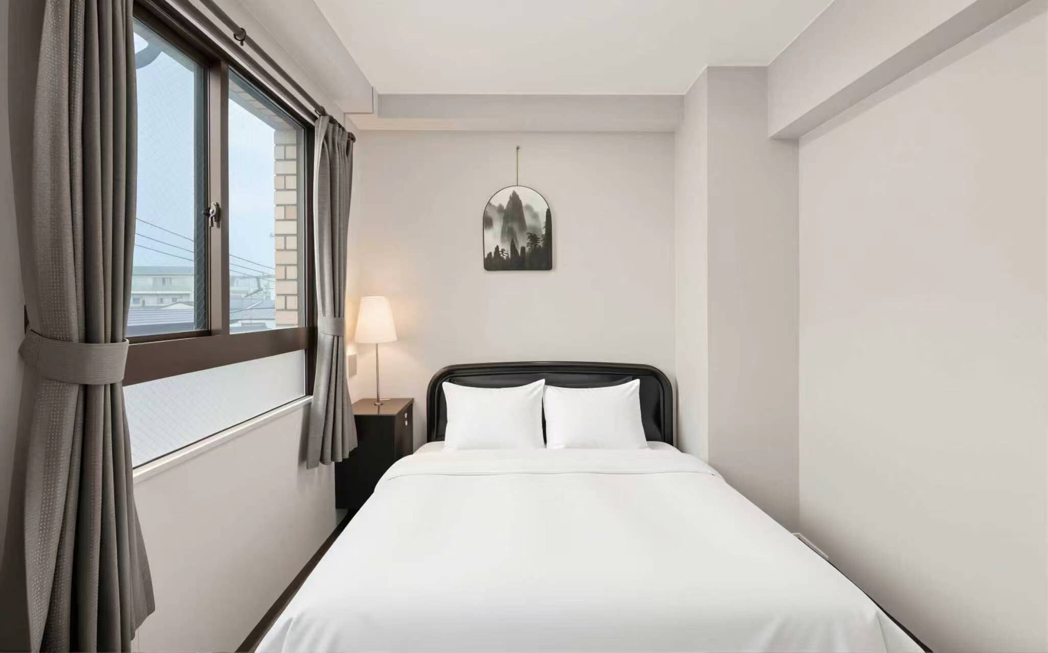 A hotel room with a bed, a nightstand, a lamp, a window with curtains, and a wall-mounted picture.