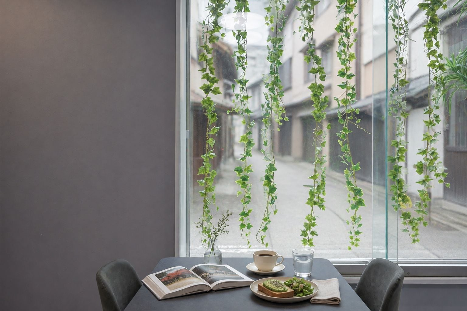 A table with a book, a cup, a glass of water, and a plate with food, positioned in front of a window with hanging plants.