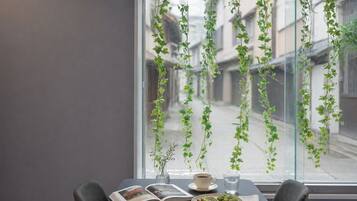 A table with a book, a cup, a glass of water, and a plate with food, positioned in front of a window with hanging plants.