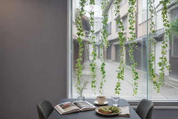 Una mesa con un libro, una taza, un vaso de agua y un plato con comida, ubicada frente a una ventana con plantas colgantes.