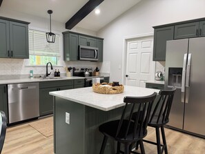A modern kitchen with a central island, dark green cabinets, stainless steel appliances, and a window with blinds. - Cozy and fun modern cottage in Luray, VA. (Luray)