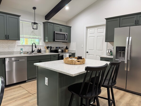 A modern kitchen with a central island, dark green cabinets, stainless steel appliances, and a window with blinds. - Cozy and fun modern cottage in Luray, VA. (Luray)