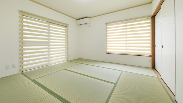 Ein Zimmer mit Tatami-Matten, zwei Fenstern mit Jalousien, einer Deckenleuchte und einer Wand-Klimaanlage.