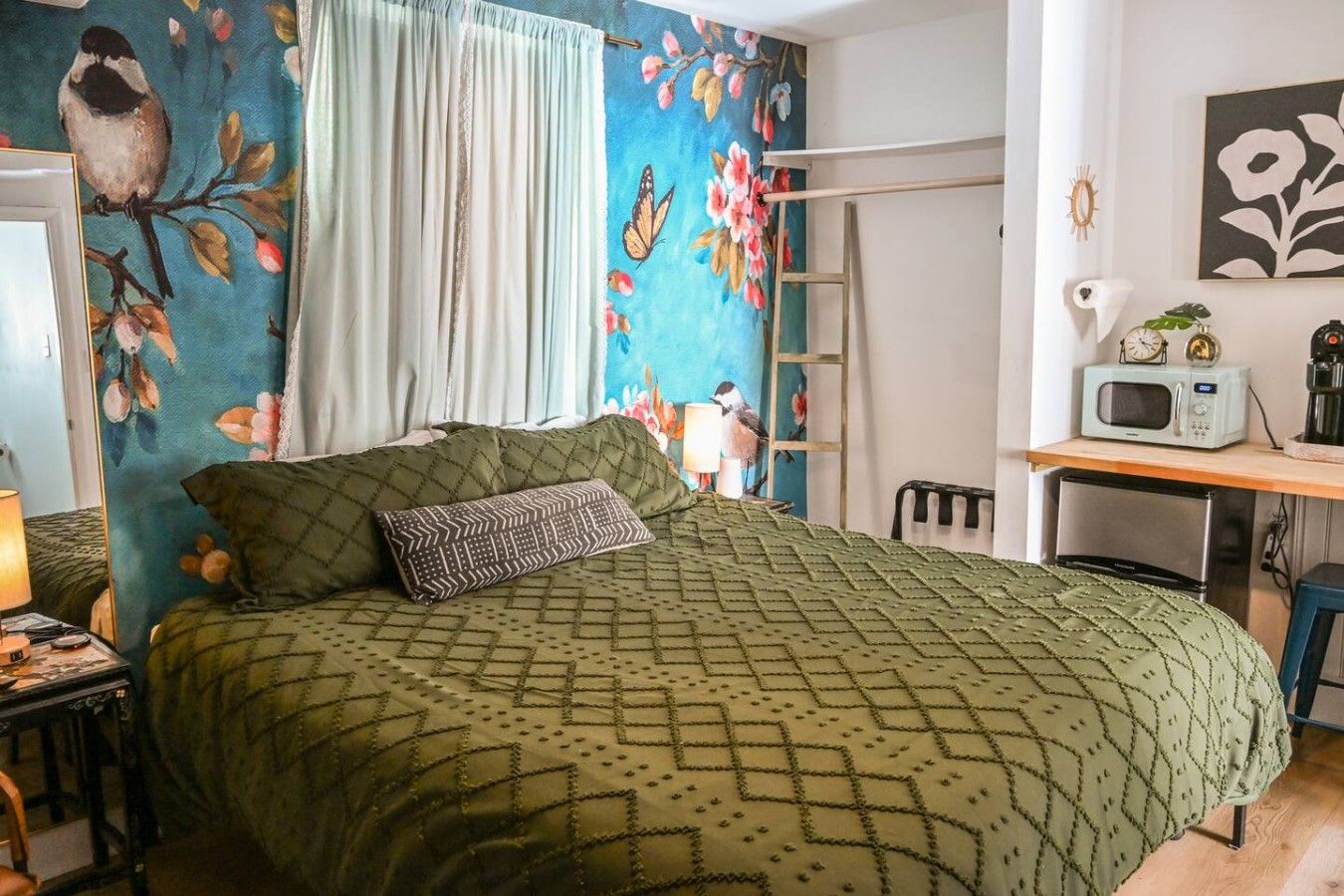 A bedroom with a bed, a desk, a chair, a lamp, and a wall with a bird and floral painting.
