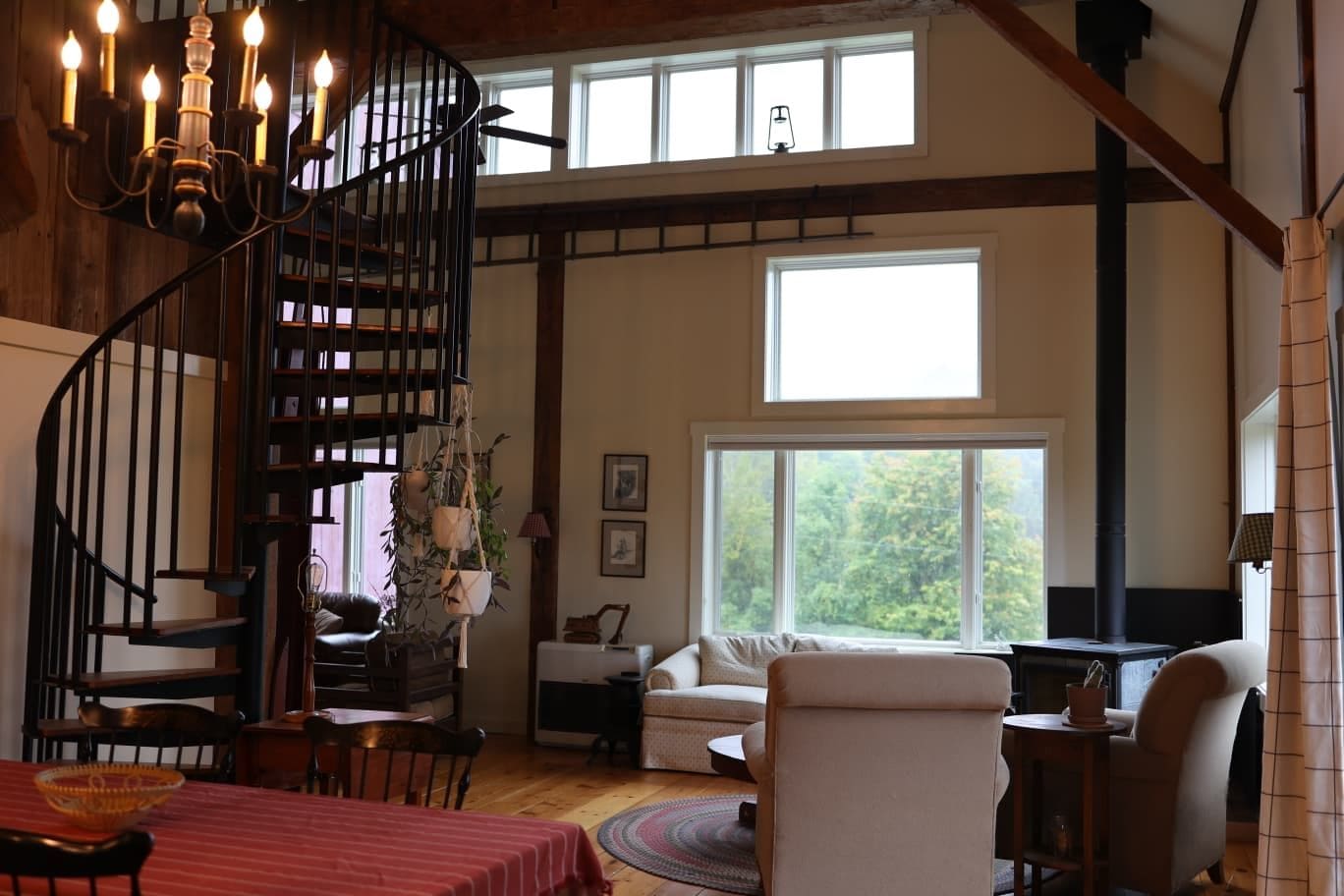 Uma escadaria em espiral iluminada por um candelabro, uma sala de jantar com toalha vermelha, uma sala de estar com sofá e lareira, e janelas amplas com vista para árvores.