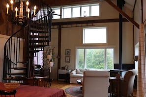 A spiral staircase with a chandelier, a dining area with a red tablecloth, a living room with a sofa and a fireplace, and large windows with a view of trees.