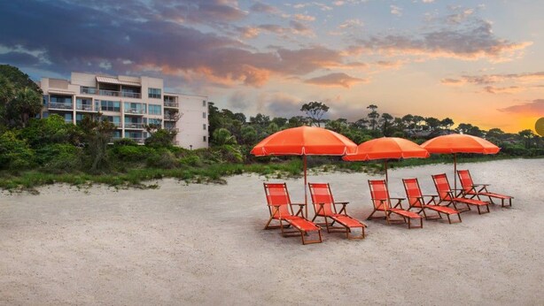 Una spiaggia con lettini e ombrelloni, un edificio sullo sfondo e un cielo al tramonto.