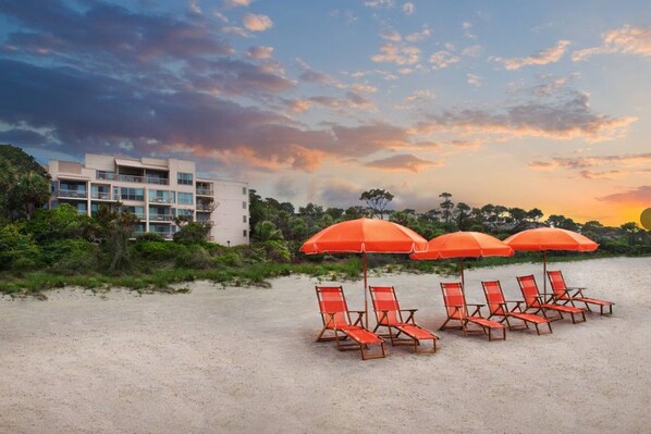 Une plage avec des chaises longues et des parasols, un bâtiment en arrière-plan et un ciel au coucher du soleil.