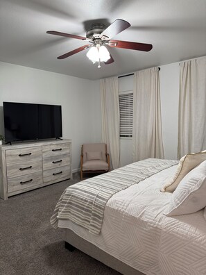 Un dormitorio con cama, televisión, sillón y ventilador de techo.