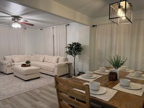 Una sala de estar moderna con sofá, reposapiés y una mesa de comedor con platos y tazones blancos.