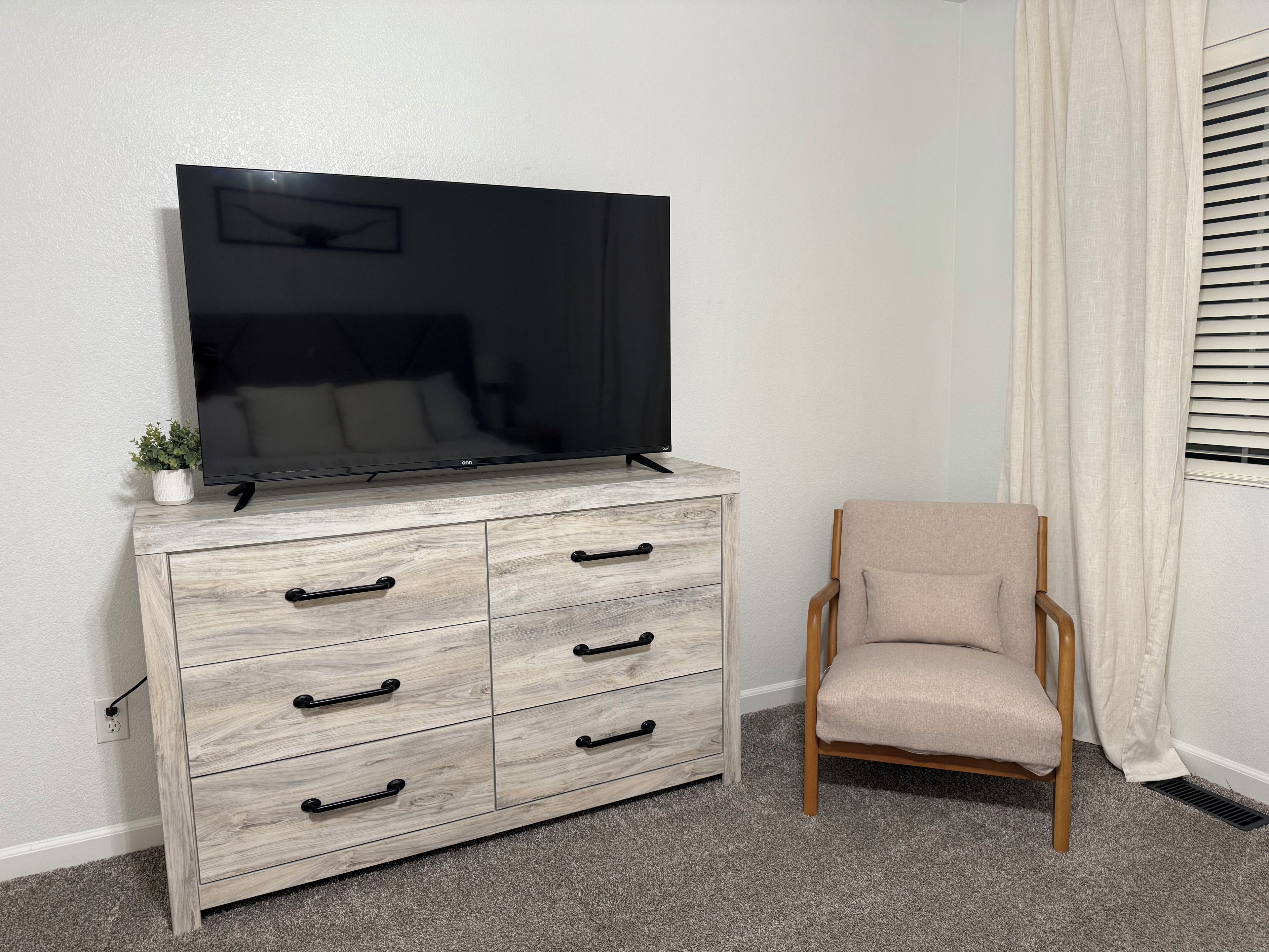 A room with a wooden dresser, a television on top, a chair, and a window with curtains.