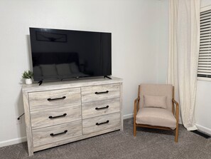 Una habitación con un aparador de madera, un televisor encima, una silla y una ventana con cortinas.