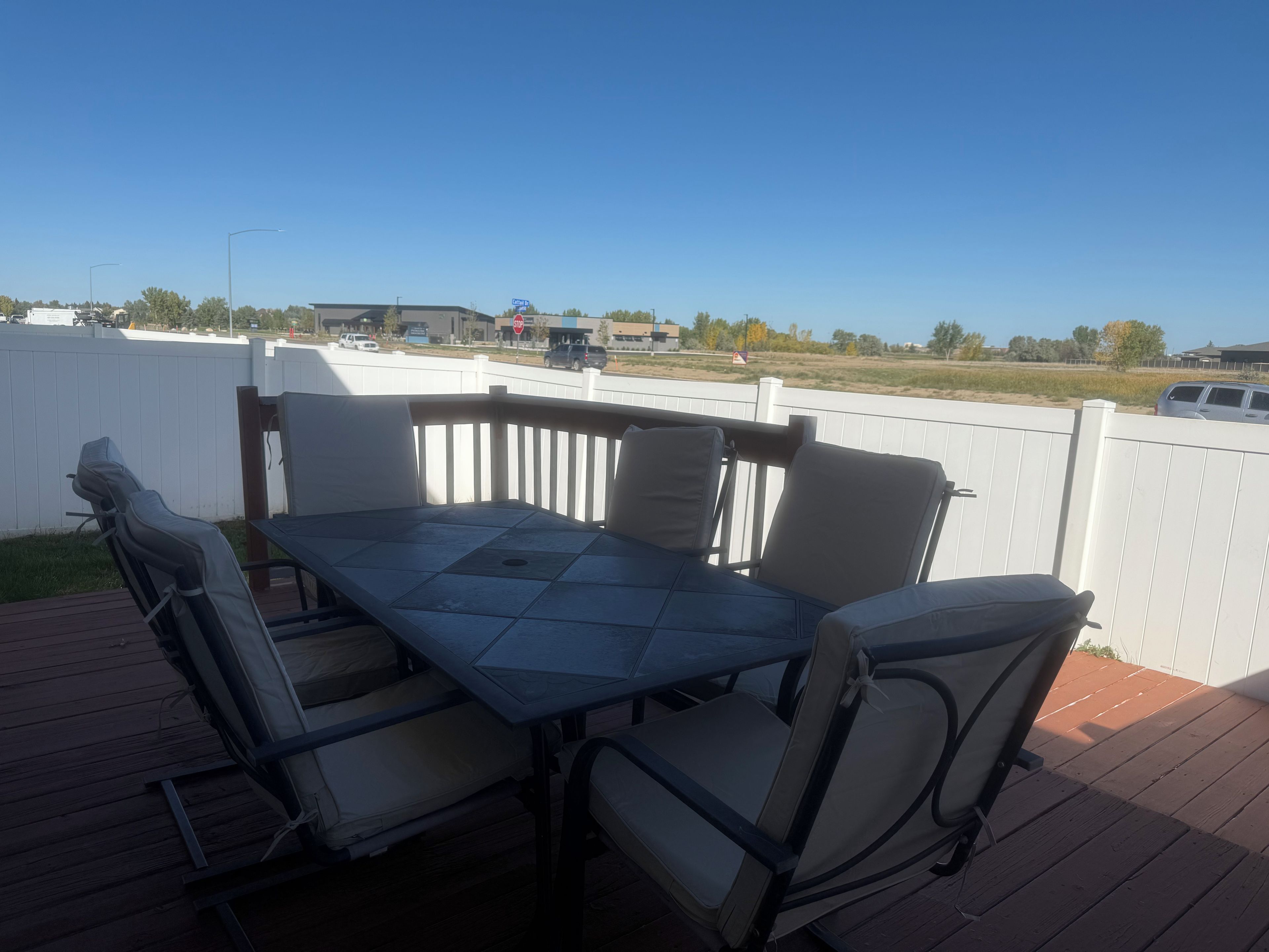 An outdoor patio with a table and chairs, a white fence, and a view of a road and buildings in the distance.